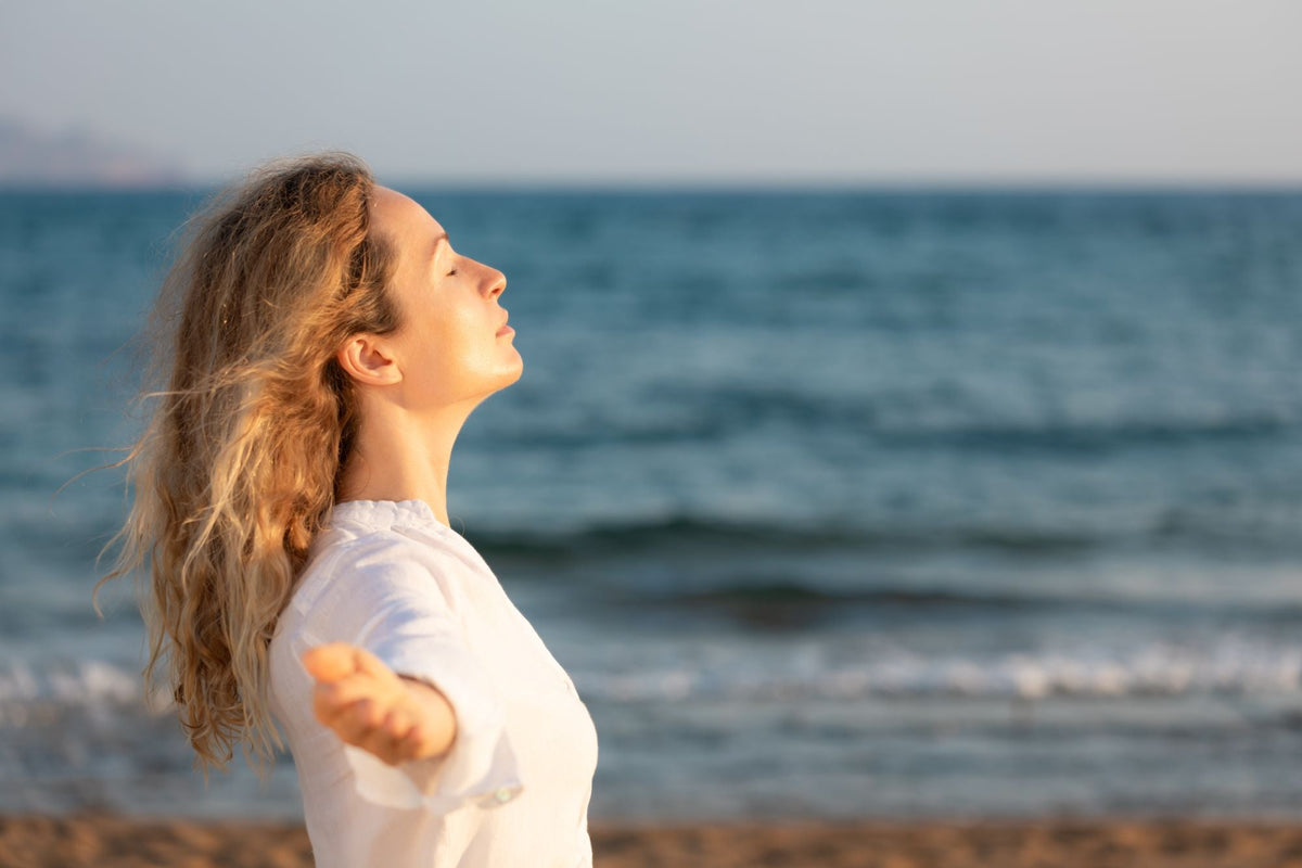 Woman standing by the ocean with eyes closed and arms outstretched, her face warmed by sunlight.